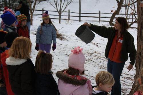 A group of children watches as a woman holds a bucket, surrounded by snow and trees in a winter setting.