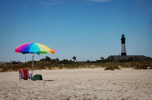 A colorful beach umbrella and chair on sandy shore, with a lighthouse in the background under a clear blue sky.