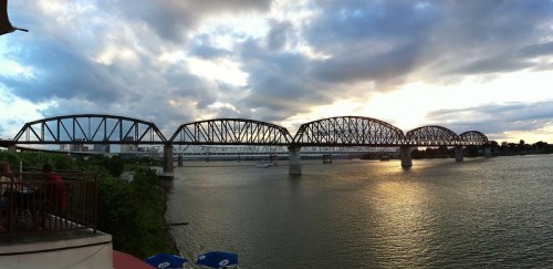 A panoramic view of a bridge over a river at sunset, with clouds and greenery in the foreground.