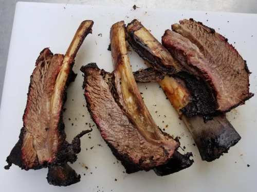 Smoked beef ribs with a charred exterior, arranged on a white cutting board.