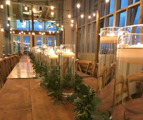A long wooden table set with glass candle holders and greenery, illuminated by string lights in a bright venue.