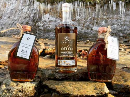 Two bottles of Starlight Distillery bourbon whiskey sit on rocky ground, with a third bottle in the center, all surrounded by ice.
