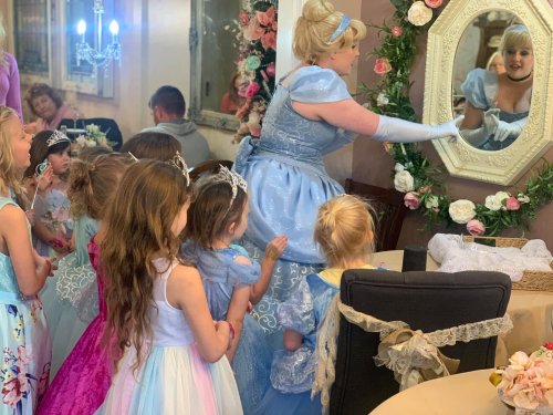 A princess in a blue gown interacts with children in fancy dresses, reflecting in a mirror at a festive gathering.