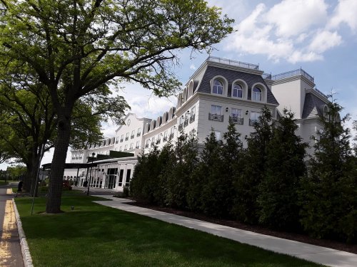 A large white hotel building with a sloped roof, surrounded by green trees and a manicured lawn under a blue sky.