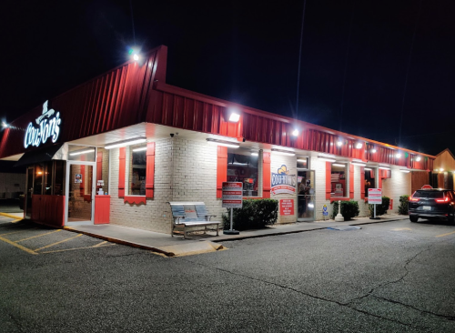 A brightly lit restaurant exterior at night, featuring red accents and signage, with parked cars nearby.