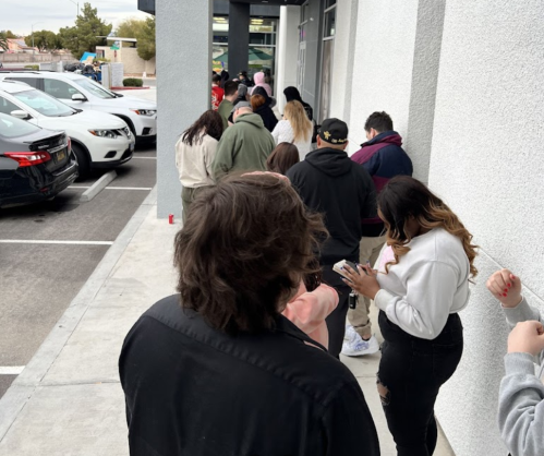 A line of people waiting outside a building, with cars parked nearby and a cloudy sky above.