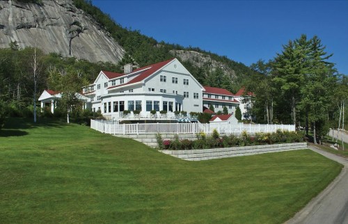 A large white building with a red roof, surrounded by green grass and trees, set against a rocky hillside.