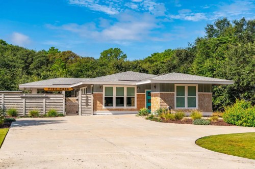Modern single-story home with a landscaped yard, large windows, and a driveway, surrounded by trees under a blue sky.