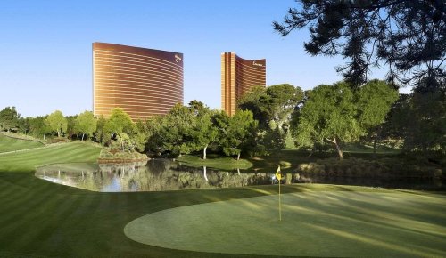 Two tall, modern hotel towers rise above a lush golf course with a pond, framed by trees under a clear blue sky.