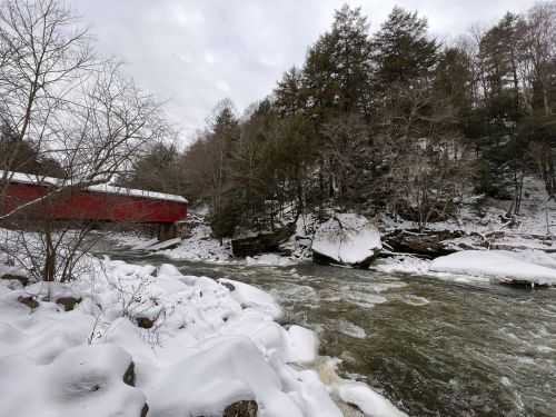 A snowy landscape featuring a river, rocky banks, and a red covered bridge surrounded by trees.