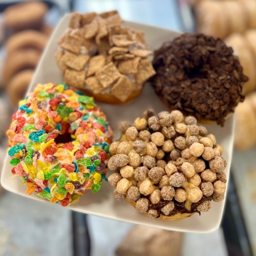 A plate of four colorful donuts topped with cereal, chocolate, and sprinkles, showcasing a variety of textures and flavors.