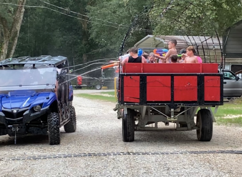 A blue off-road vehicle sprays water at a group of people on a red trailer, enjoying a fun water fight outdoors.