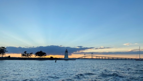 Sunset over water with a lighthouse and bridge in the background, surrounded by trees and clouds.