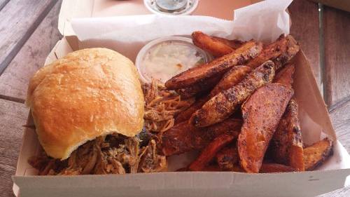 A takeout box with a pulled pork sandwich and a side of crispy sweet potato fries.
