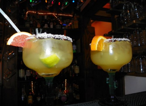 Two colorful margaritas with fruit garnishes, served in large glasses, against a backdrop of a well-stocked bar.