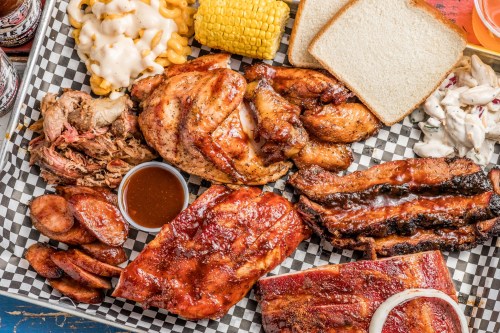 A vibrant platter of barbecue featuring pulled pork, chicken, ribs, sausage, mac and cheese, corn, and bread.
