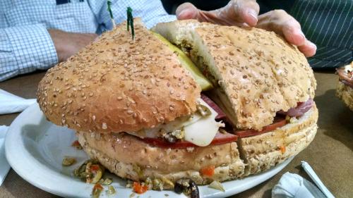 A large sesame seed bun sandwich filled with layers of meats, cheese, and pickles, served on a plate.