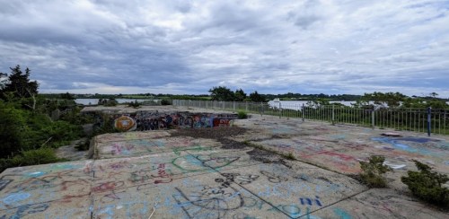 A graffiti-covered concrete platform overlooks a cloudy sky and distant landscape with trees and water.