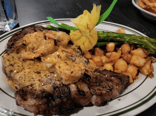 A plate featuring a grilled steak topped with shrimp, asparagus, and seasoned potatoes, garnished with a yellow ribbon.
