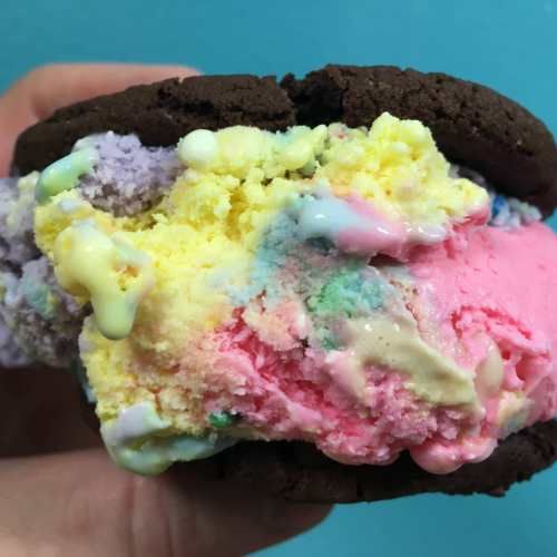 A hand holding a colorful ice cream sandwich with pastel ice cream between two chocolate cookies.