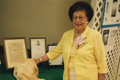 An elderly woman in a yellow jacket smiles beside a display of framed documents and a floral arrangement.