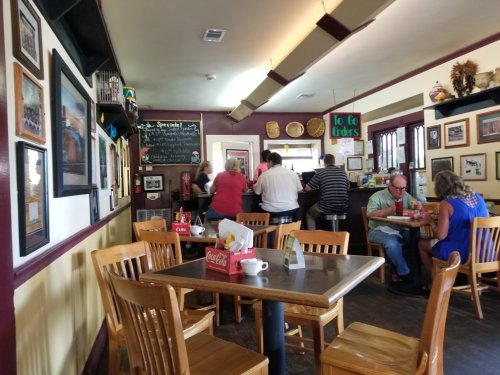 A cozy restaurant interior with wooden tables, patrons at the counter, and walls adorned with framed photos.