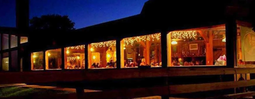 A warmly lit restaurant with large windows, showcasing diners inside against a twilight sky.