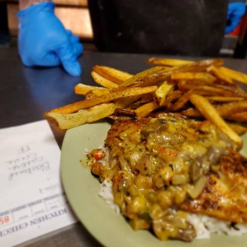 A plate of grilled chicken with sauce over rice, served with golden fries, and a gloved hand in the background.