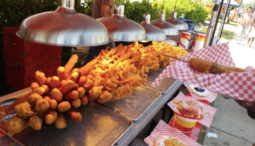 A food stall displaying a variety of fried items, including corn dogs and fried chicken, under bright metal lamps.