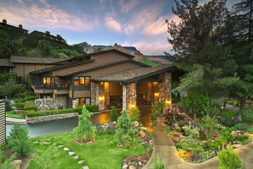 A scenic lodge surrounded by lush greenery and mountains, featuring a stone facade and a welcoming entrance.