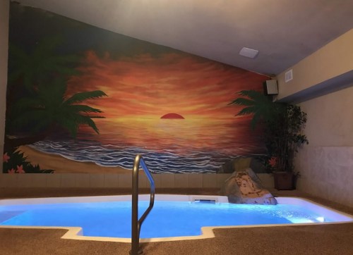 Indoor pool area with a sunset mural, palm trees, and a rock feature beside the water.