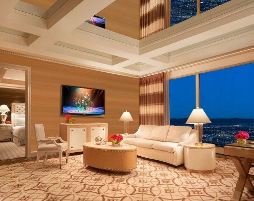Luxurious hotel suite with a modern design, featuring a sofa, TV, and stunning city views through large windows.