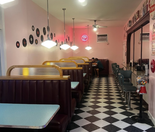 A retro diner with pink walls, checkered floor, and booths, featuring vinyl records and a gumball machine.