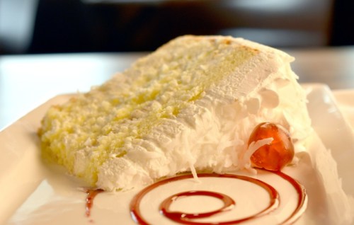A slice of fluffy white cake topped with whipped cream, a cherry, and a swirl of red sauce on a white plate.