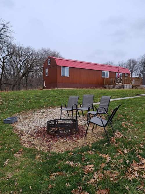 A fire pit surrounded by four chairs, with a cabin in the background and trees in a grassy area.
