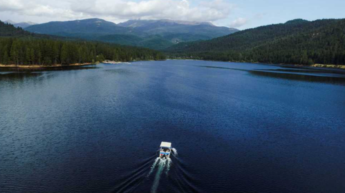 A boat glides across a serene lake surrounded by lush forests and mountains under a partly cloudy sky.