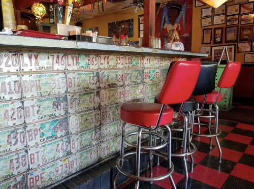 A colorful bar with vintage license plates on the wall and red and black stools in a lively setting.