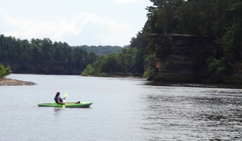 A person kayaking on a calm river surrounded by lush greenery and rocky cliffs in the background.