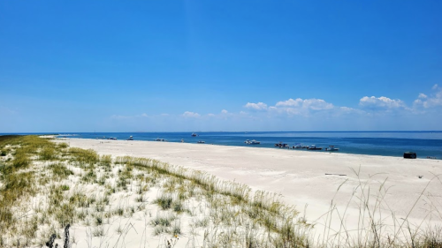 A serene beach scene with white sand, gentle waves, and boats in the distance under a clear blue sky.