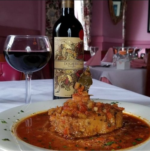 A plate of savory meat stew garnished with herbs, accompanied by a glass of red wine and a bottle on a table.