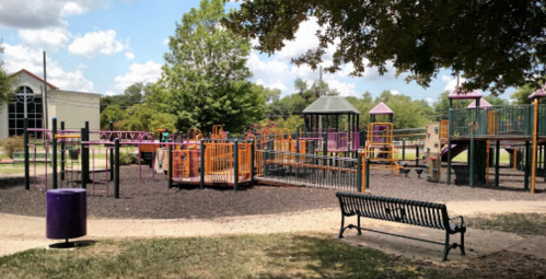 Colorful playground with slides and climbing structures, surrounded by trees and a bench in a sunny park setting.