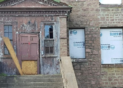 Weathered building with a wooden door, boarded windows, and construction materials covering the facade.