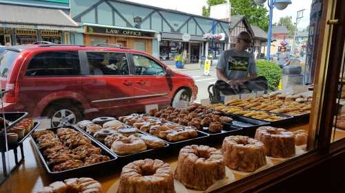 A display of various baked goods in a shop window, with a red van parked outside and a person browsing nearby.