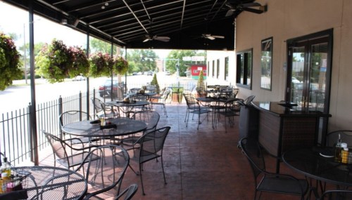 Outdoor dining area with black metal tables and chairs, surrounded by hanging plants and a view of the street.
