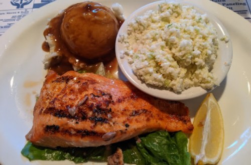 Grilled salmon fillet on lettuce, served with mashed potatoes, coleslaw, and a lemon wedge.