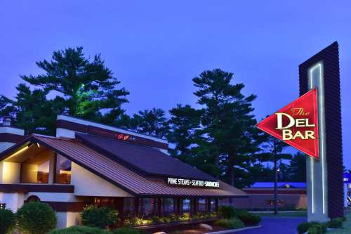 Exterior of The Del Bar restaurant at dusk, featuring a modern sign and surrounded by trees.