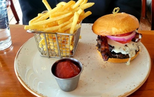 A plate with a burger topped with bacon, onions, and sauce, alongside a basket of fries and a small cup of ketchup.