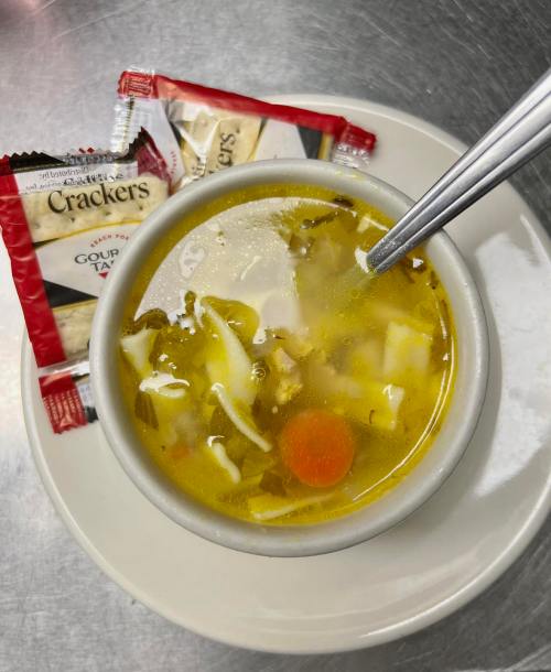 A bowl of soup with vegetables and noodles, accompanied by two packets of crackers on a plate.
