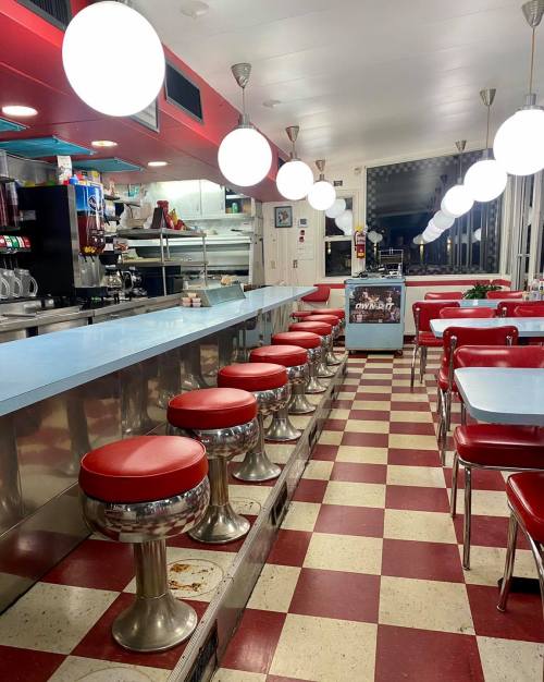 A retro diner interior featuring red and white checkered floors, chrome stools, and round pendant lights.