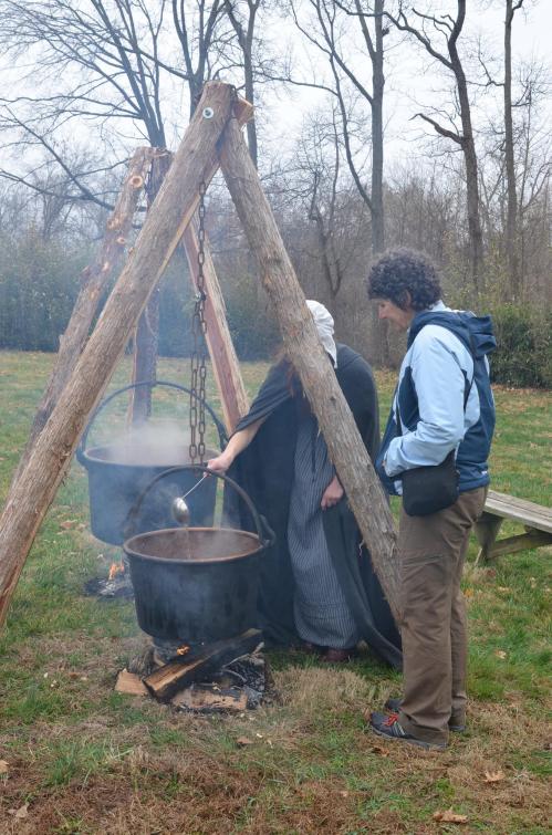 A person in a cloak stirs a pot over an open fire, while another person observes nearby in a wooded area.